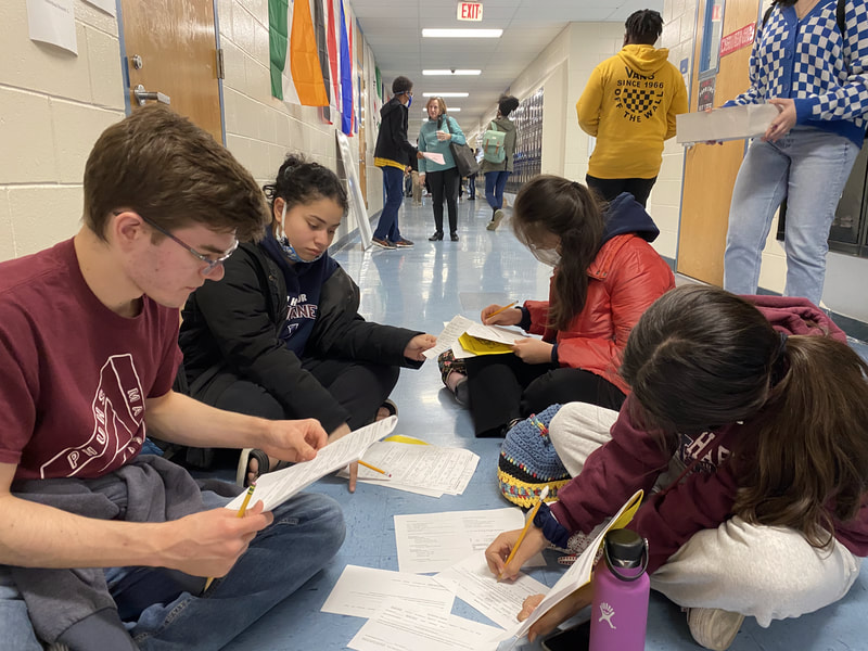 A group of students sit on the floor while leafing through math test packets. 